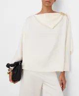 Top fluido satinado color marfil, con línea rectangular, cuello pañuelo que gira desde el delantero hacia la espalda sujeto con hebilla decorativa dorada en el hombro izquierdo y manga larga con dos botones en el puño.