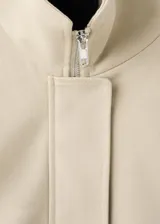 Chaqueta oversize de algodón color beige, con cuello alto, cierre frontal oculto con cremallera, mangas largas con puños abotonados, bolsillos laterales y bajo fruncido elástico.