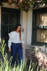 Camisa de color natural con cuello redondo, mangas largas con puños y detalle de volados amplios en la parte superior delantera y hombros. Presenta cierre frontal con botones.