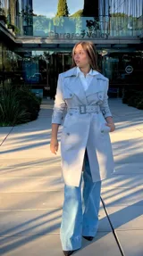 Trench coat gris de corte clásico con doble botonadura, solapas amplias, cinturón ajustable con hebilla y mangas con trabillas en los puños.