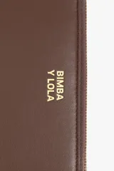 Billetera rígida de color negro con cierre de cremallera perimetral. Presenta el logo metálico de Bimba y Lola en color dorado en el frente y un tirador de cordón trenzado en tonos marrón y amarillo. El interior cuenta con compartimentos para tarjetas y un monedero con cierre.