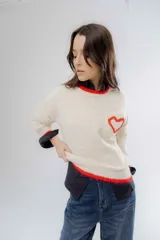 Buzo de punto color crema con detalles en rojo en el cuello, puños y ruedo. Presenta un bolsillo frontal en el pecho con un corazón bordado en relieve.