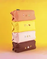 Neceser rectangular de tela color ladrillo con cierre superior y tres bordados pequeños: una hoja de arce, una taza de té y un croissant.