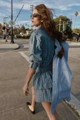 Vestido corto de manga larga con cuello solapa y cierre frontal de botones a presión. Presenta un diseño a cuadros en tonos azul y marrón, cintura ajustable con cinturón de hebilla y falda con volantes en el bajo.