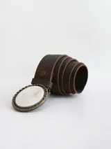 Cinto de cuero color chocolate oscuro con forro de cuero. Presenta una hebilla circular con borde metálico labrado y centro de guampa, con sistema de broche suizo intercambiable.