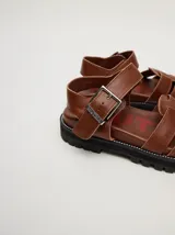 Sandalias tipo romanas de cuero vacuno color beige, con diseño de tiras entrelazadas, hebilla metálica lateral y plataforma de goma dentada.