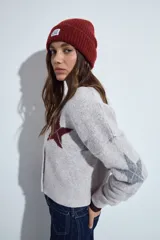 Cardigan corto tejido en color gris, con escote en V y cierre frontal de botones. Presenta un diseño estampado con estrellas y líneas geométricas en tonos oscuros y rojizos, con mangas largas y puños acanalados.