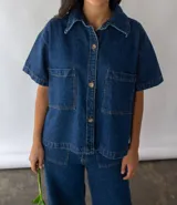 Camisa de denim azul de algodón reciclado, corte amplio, con cuello clásico, manga corta, cierre frontal con botones y dos bolsillos de parche en el pecho.