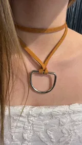 Gargantilla tipo choker confeccionada en tira de gamuza color tostado, con un dije metálico en forma de semicírculo plateado.