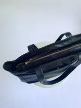 Cartera tipo tote de cuero graneado negro, con diseño clásico y textura uniforme. Cuenta con doble asa de hombro, bolsillo frontal con cierre magnético, bolsillo trasero con cierre metálico y compartimento principal con cierre. El interior está forrado y dispone de múltiples bolsillos organizadores.