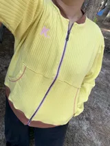Campera de algodón premium color amarillo con cuello, puños y dobladillo en marrón. Tiene cierre frontal, bolsillos delanteros con ribetes rosados y la letra 'K' bordada en rosa en el pecho.