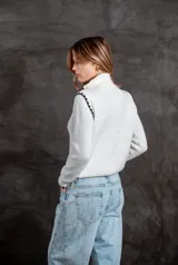 Buzo de punto color blanco con cuello polo y cierre frontal. Presenta detalles de rayas negras en los hombros y puños, con un diseño de tejido acanalado.