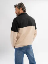 Buzo de felpa con diseño color block, combinando negro en la parte superior y beige en la parte inferior. Presenta cuello alto con cierre frontal parcial y puños elásticos.