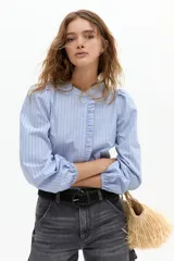 Camisa de algodón con estampado de rayas verticales en tonos azul y blanco. Presenta cuello mao, cierre frontal oculto con detalle de volado lateral y mangas abullonadas con puños elásticos.
