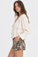 Blusa de seda color marfil con escote en V, mangas largas con puños fruncidos y múltiples volados en capas adornados con tachas metálicas.