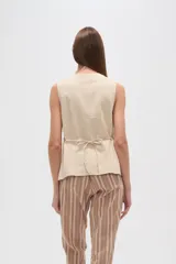 Chaleco color beige claro, sin mangas, con escote en V profundo y cierre frontal asimétrico con botones.