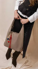 Cartera tipo hobo de cuero sintético color suela, con interior espacioso y costuras decorativas frontales. Incluye asa de hombro fija, monedero rectangular colgante a juego y un llavero amuleto con cuentas de madera, ojos turcos y detalles en rojo.