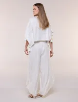 Pantalón palazzo cruzado color blanco con trama en hilo metalizado lurex, cintura elastizada con cordón ajustable con borlas y diseño de piernas anchas con bordes de puntilla y tajo profundo en el frente.