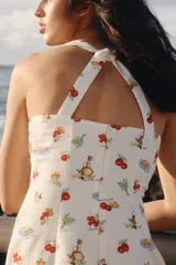 Vestido mini de color beige con un estampado de tomates, aceitunas y botellas de aceite, y un borde inferior rojo con ilustraciones de una sombrilla de playa y vegetales. Cuenta con escote cuadrado y tirantes anchos cruzados en la espalda.