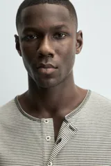 Camiseta de manga larga con cuello panadero (henley) y cierre de tres botones. Presenta un patrón de rayas horizontales finas en tonos beige y negro, con pespuntes a contraste en el cuello, puños y bajo.