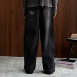 Pantalón de jean negro con corte baggy, de tiro medio y pierna ancha. Presenta un parche de cuero naranja en la cintura trasera y un bolsillo trasero con un parche bordado de la marca Nude Project. El ruedo tiene un acabado deshilachado.