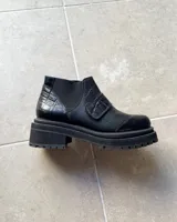 Botineta de cuero vacuno color negro con suela track de goma. Presenta una combinación de texturas en el cuero, incluyendo paneles con efecto croco en el talón y en la tira decorativa sobre el empeine, además de paneles laterales elásticos.