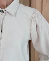 Camisa de color arena con mangas abullonadas, puños anchos con botones y cuello clásico. Presenta un diseño de corte recto con costuras visibles y cierre frontal oculto.