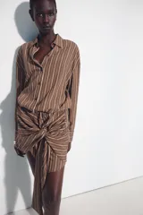 Vestido corto marrón con rayas verticales blancas, cuello camisero, manga larga y detalle de nudo y lazada en la falda. Cuenta con cierre frontal de botones.