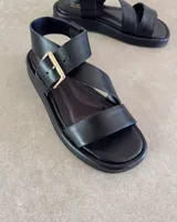 Sandalia de cuero negro con suela plana, tiras anchas cruzadas y cierre con hebilla metálica dorada en el tobillo.