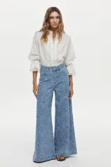 Camisa blanca de algodón con cuello alto, detalles de encaje broderie en el frente, mangas abullonadas y puños con volados y encaje. Cierre frontal con botones tapados.