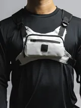 Bolso de pecho táctico color gris claro con correas ajustables negras y hebillas. Cuenta con tres compartimentos: uno trasero de red y dos frontales con cierre, uno de ellos con un parche negro con logo.