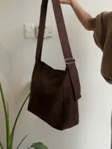 Bolso tipo bandolera de tamaño mediano, confeccionado en material con acabado efecto gamuza color chocolate. Presenta un diseño minimalista con cierre superior de cremallera y correa larga regulable con herrajes metálicos.
