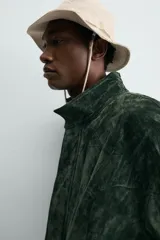 Cazadora regular fit de piel con acabado en ante color verde oscuro, de la colección especial Gabriel Azevedo x ZARA. Cuenta con cuello subido, manga larga, bolsillos de vivo en la cadera, bolsillo interior y bajo acabado con elástico. Cierre frontal con cremallera oculta por solapa con botones a presión.