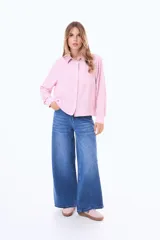 Camisa blanca de manga larga con cuello clásico y cierre frontal de botones negros en contraste. Presenta un bolsillo frontal en el pecho y un diseño de líneas simples y corte recto.