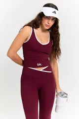 Conjunto deportivo compuesto por un top y un pantalón largo en color bordó. El top presenta un diseño con cuello redondo y ribetes blancos, con el logo 'bk.' estampado en el frente. El pantalón es de corte ajustado con cintura cruzada en V y detalle de ribete blanco, con terminación acampanada en los tobillos.