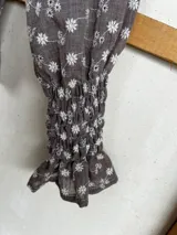 Blusa de algodón con diseño de nido de abeja en el frente y la espalda, cuello alto con volado y mangas largas con puños elásticos terminados en volado. Presenta un estampado floral pequeño en tonos marrón y crudo.