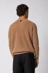 Sweater de tejido de punto grueso color tostado, con cuello redondo y mangas largas con puño elástico que incluye una etiqueta decorativa.