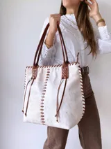 Cartera tipo tote de cuero vacuno con acabado texturizado en tono blanco hueso. Presenta detalles de costuras artesanales con tientos de cuero marrón en los bordes y correas de hombro a juego.