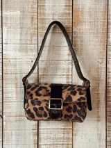 Cartera tipo bandolera con solapa y estampado animal print de leopardo, con detalle de tira y hebilla metálica frontal en color negro. Cuenta con asa corta ajustable con ojales metálicos.