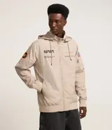 Campera rompevientos color beige con capucha, cierre frontal y bolsillos laterales. Presenta estampado de la NASA en el pecho, parche circular en la manga derecha y bandera de Estados Unidos en la manga izquierda.
