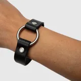 Pulsera de cuero sintético negro con gran anillo metálico plateado en el centro y tachas a presión plateadas en los extremos.