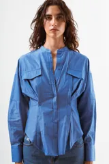 Camisa azul de algodón con finas rayas blancas verticales, cuello mao, manga larga y detalle de pinzas y botones en la zona de la cintura. Posee dos bolsillos de parche con tapa en el pecho.