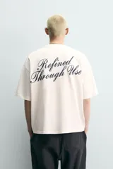 Camiseta blanca de corte relajado, con cuello redondo, manga corta y texto estampado en contraste en el pecho.