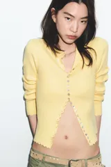 Chaqueta de punto acanalado en color blanco, con cuello redondo, manga larga y cierre frontal con botones nacarados. Presenta terminaciones con ondas en el ruedo y los puños.