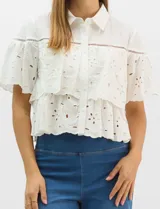 Camisa de algodón blanco con diseño romántico, cuello clásico y cierre frontal de botones. Presenta paneles de tejido bordado suizo con motivos calados, capas superpuestas con volados y mangas cortas con silueta acampanada.