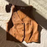 Chaleco de cuero sintético metalizado en color dorado, con escote en V, cierre frontal de botones y detalles de tachas metálicas en los bordes.