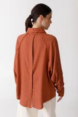 Camisa color ladrillo con rayas verticales finas color crema, de corte recto y mangas largas con puños abotonados. Presenta cuello camisero, cierre frontal con botones y detalle de flecos en los hombros.