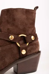 Bota corta de gamuza color marrón, con taco cuadrado y punta fina. Presenta una correa con tachas doradas y una argolla metálica decorativa alrededor del tobillo. Cuenta con cierre lateral dorado.