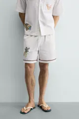 Bermuda de corte relajado confeccionada en algodón, color blanco con vivo en contraste color marrón en el ruedo. Presenta cintura elástica con cordón ajustable y un bordado decorativo en la pierna derecha que incluye una palmera y un gallo.