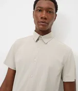 Camisa de manga corta color beige, con cuello camisero, cierre frontal de botones y diseño liso.